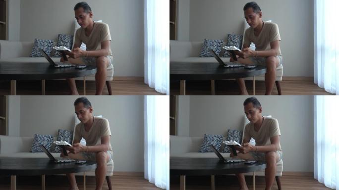 亚洲男人坐在客厅的教练时使用笔记本电脑。在家工作，带着家庭的相框