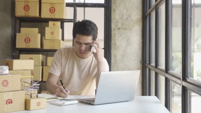 穿着t恤的年轻亚洲商人在家庭办公室用笔记本电脑接受订单的包裹中工作，名字看起来像是在做包装箱交付，中