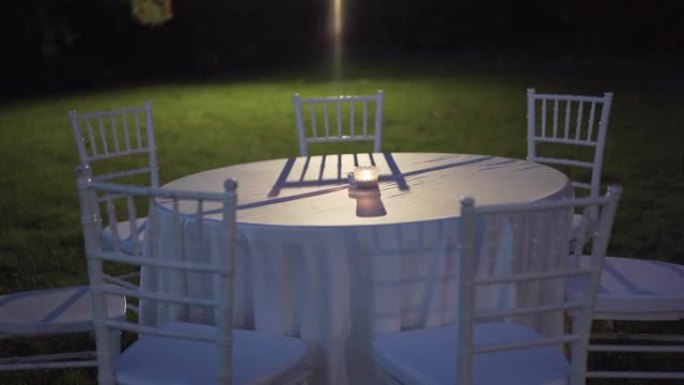 晚上在花园里铺着桌布和白色椅子的圆桌，桌子中央燃烧着小蜡烛，背景照明桌子设置上的聚光灯，空旷的花园，