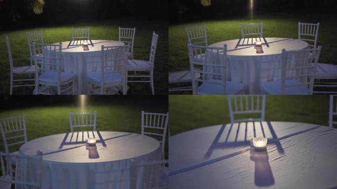 晚上在花园里铺着桌布和白色椅子的圆桌，桌子中央燃烧着小蜡烛，背景照明桌子设置上的聚光灯，空旷的花园，