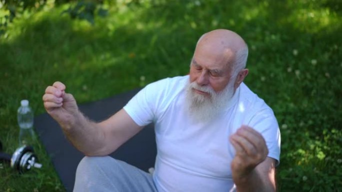 高角视图的老高加索大胡子男人在莲花姿势冥想坐在运动垫上，以慢动作闭上眼睛。平静自信的高级瑜伽士在夏季