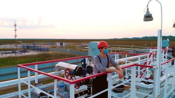 一名戴着头盔和口罩的石油和天然气工业经营者在日落时站在压力容器上。石油工人在日出时在油气田工作。炼油