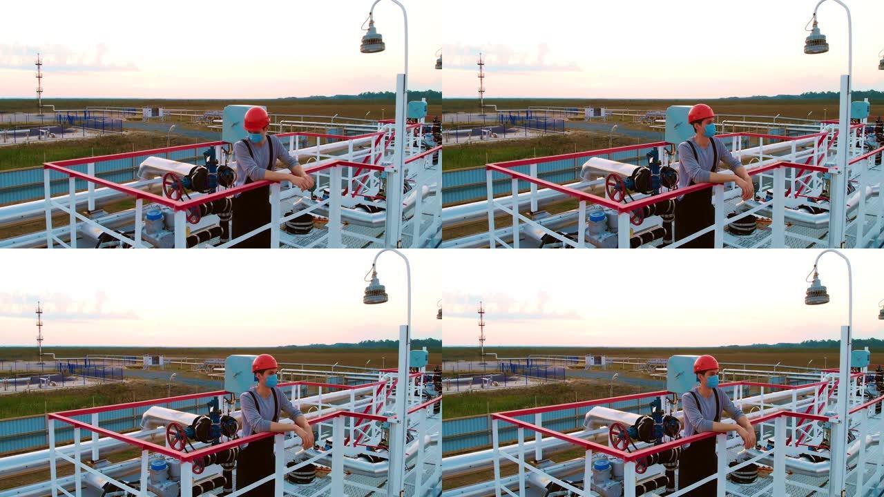 一名戴着头盔和口罩的石油和天然气工业经营者在日落时站在压力容器上。石油工人在日出时在油气田工作。炼油