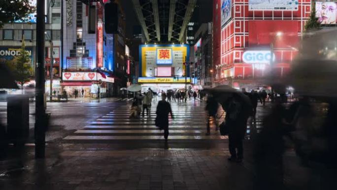 时间流逝在雨夜，人们在日本东京秋叶原电气镇使用雨伞步行横穿道路，汽车交通运输。动漫，游戏，娱乐区。亚