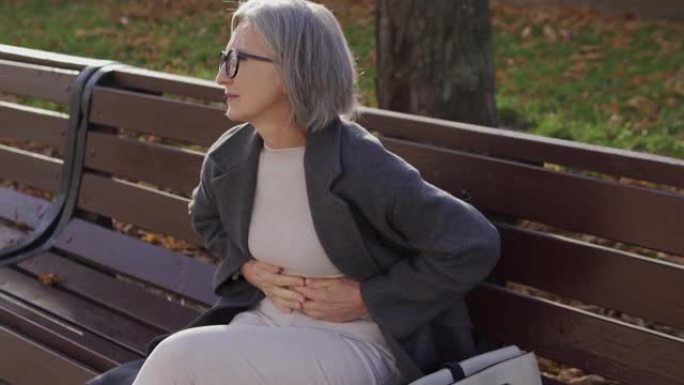 优雅的高级女性坐在公园的长凳上，痛苦地皱着眉头，感到急性腹痛，患有胃炎或十二指肠溃疡，阑尾破裂或胆囊