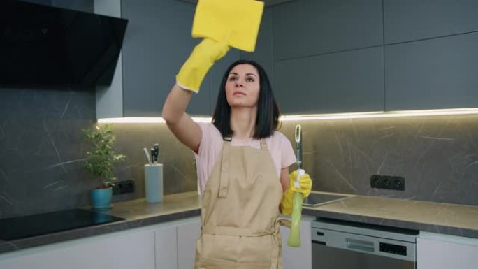 在厨房的家中，穿着黄色橡胶手套和围裙用洗涤剂洗涤窗的高加索家庭主妇的肖像。