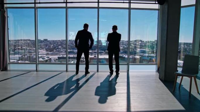 穿着深色西装的商人在休息时说话。办公室工作人员站在大窗户附近，看着城市。后视图。