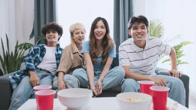 一群快乐的亚洲年轻人在家里一起看电视，吃爆米花零食，笑着一起笑。有趣的周末休闲活动，亲密的朋友关系或
