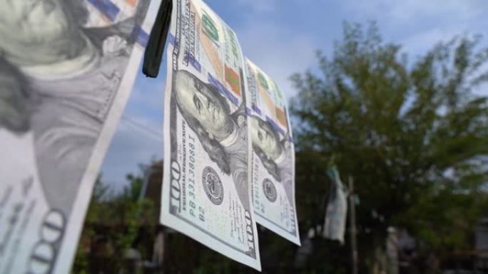 近距离可视化的现金美元洗钱。在模糊的蓝天背景下，摄像机沿着用衣夹固定的晾衣绳上的百元美钞平滑移动