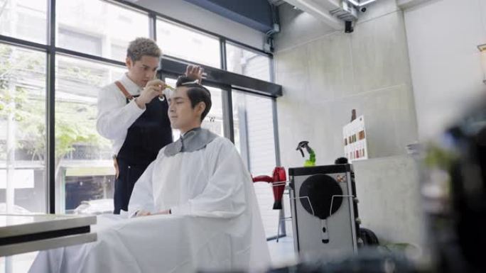 亚洲专业男性发型师穿着黑色围裙梳头，在沙龙里用剪刀剪发和修剪年轻顾客的头发。坐在商店里等着的那个人，