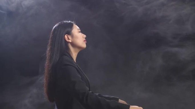 亚洲指挥家妇女的侧视图，她拿着指挥棒闭上眼睛，在迷雾的黑色工作室里展示手势