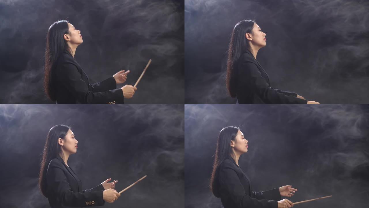 亚洲指挥家妇女的侧视图，她拿着指挥棒闭上眼睛，在迷雾的黑色工作室里展示手势