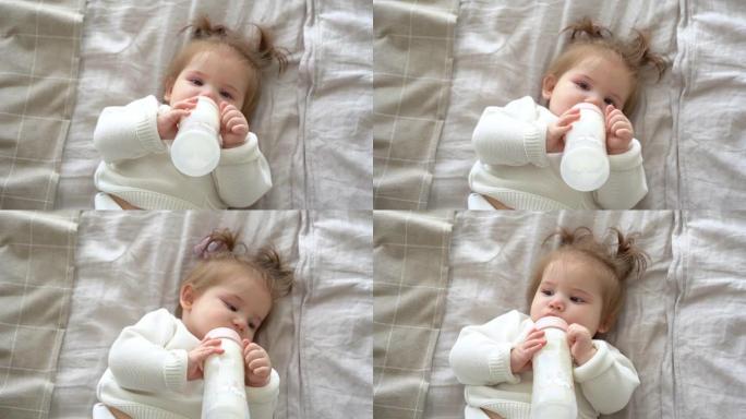 小女孩在家中从奶瓶中吸奶，肖像婴儿进食，喝奶粉婴儿配方奶粉。可爱的孩子躺在床上，穿着针织毛衣，辫子，