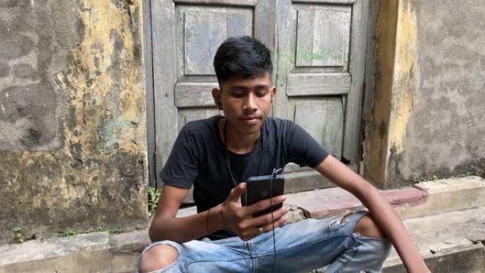一个穿着黑色t恤的年轻印度男孩坐在一栋带耳机的老建筑的台阶前