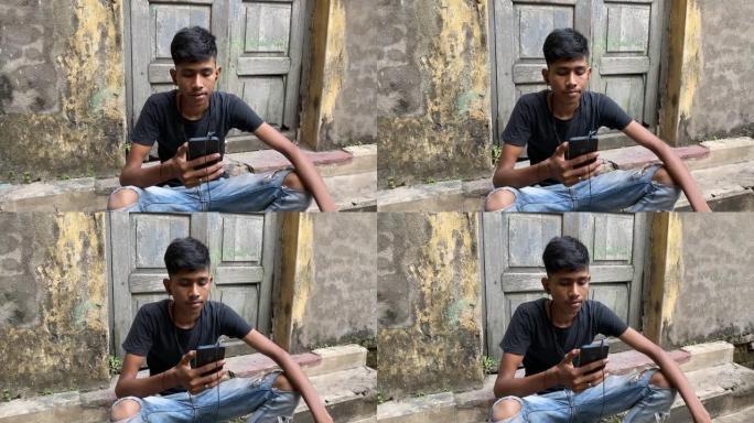 一个穿着黑色t恤的年轻印度男孩坐在一栋带耳机的老建筑的台阶前