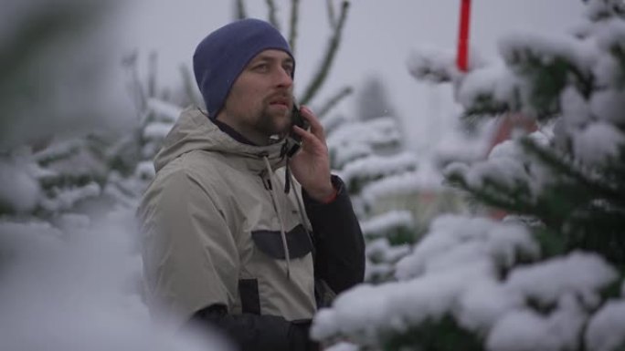 男人在圣诞节市场上选择圣诞树，并在下雪的天气中通过家人的电话获取正确选择的提示。在当地农贸市场挑选枞