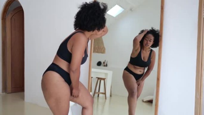 穿着黑色内衣的非洲亚裔妇女在镜子前检查自己的橘皮组织，无法相信自己的身体变得如此肥胖。世界各地吃快餐