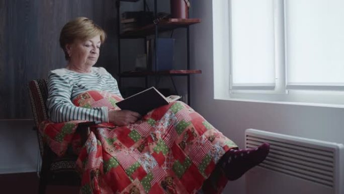 一位老妇人正盖着毯子在窗前看书。在电暖器的背景上，腿穿着温暖的袜子。寒冷的季节。节约电能。能源危机。