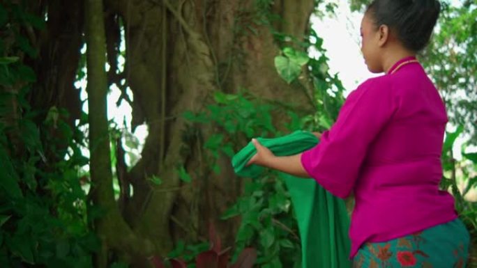 一名亚洲妇女在森林内的大树前举行仪式后，穿着传统的粉红色连衣裙穿着绿色围巾