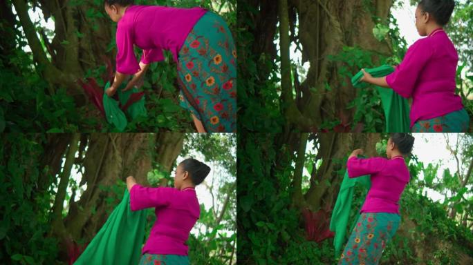 一名亚洲妇女在森林内的大树前举行仪式后，穿着传统的粉红色连衣裙穿着绿色围巾