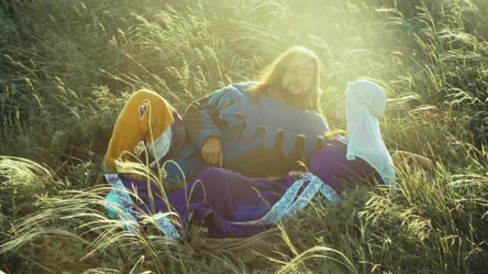 穿着中世纪服装的男人和女人的慢动作在夕阳的明亮光线中躺在高高的草丛中休息。历史镜头的概念。博客