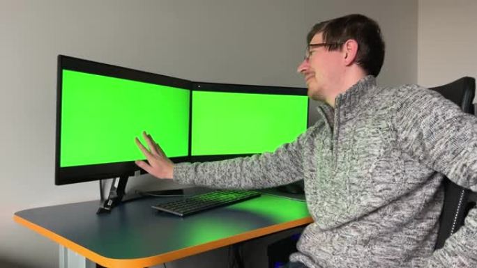 年轻的程序员艺术家网页设计师看着有两个绿色屏幕的显示器chromakey他用手轻轻地吞下其中一个显示
