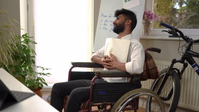 英俊的中东聪明的年轻人读拥抱书，看着微笑，坐在轮椅上在室内。积极满意的自由职业者在家庭办公室享受爱好