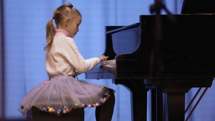 可爱的7岁女孩在彩色拱腹的灯光下在舞台上演奏钢琴。在音乐学校度过休闲的一天
