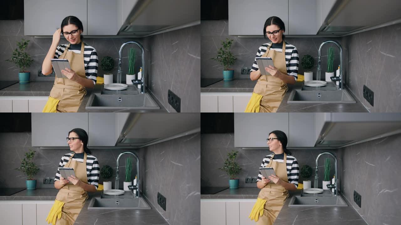 穿着眼镜的美丽年轻女子在厨房洗碗时看着数字平板电脑屏幕，例行公事，生活方式