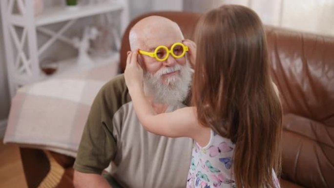 大胡子的高级白发祖父坐在沙发上，孙女戴上黄色玩具眼镜。快乐的高加索老人拥抱迷人的女孩，在室内享受休闲