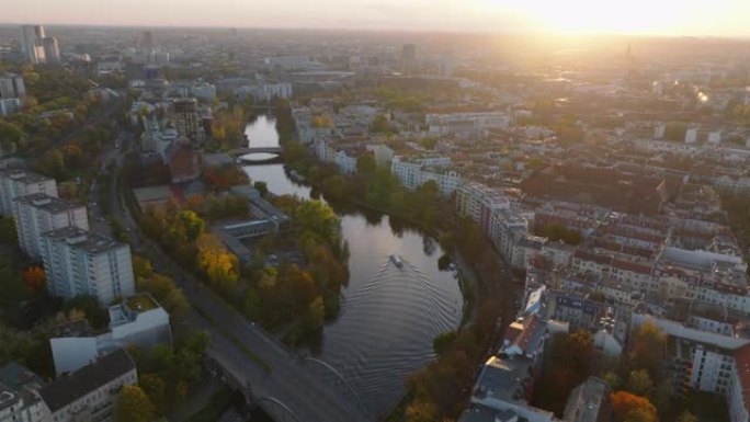 日落时分，河流蜿蜒穿过市区的风景如画的航拍画面。船在平静的水面上航行，岸边被秋天的彩树包围。德国柏林