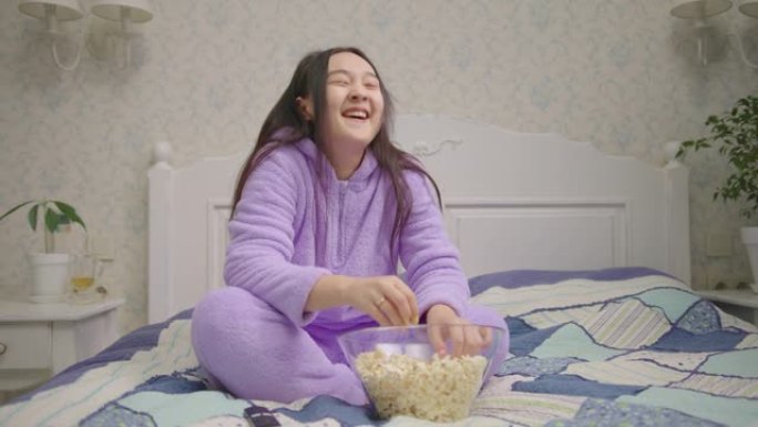 亚洲女人看搞笑电影或电视节目，独自坐在床上吃爆米花。穿着紫色睡衣的女士在电视前大笑。