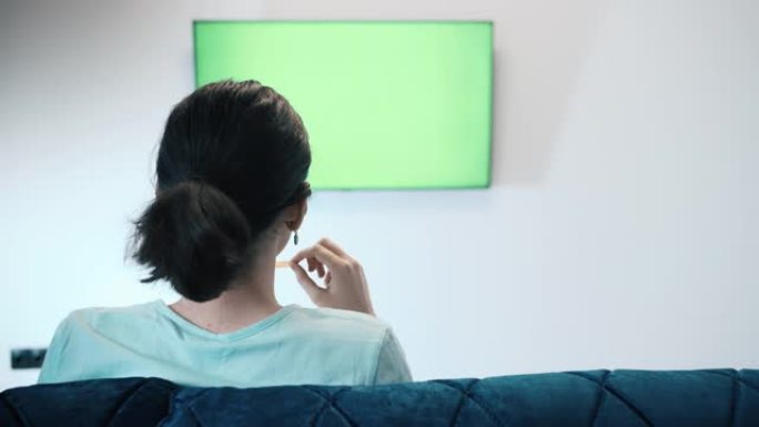 戴着帽子的黑发女孩坐在吉恩斯林电视的字体里，切换频道，绿色色度键屏幕电视，在沙发上放松，专心观看电视