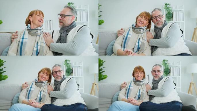 白人老年夫妇的肖像坐在客厅的沙发上。爱年长的成熟祖母和祖父互相拥抱，一起享受退休生活，然后在家里看相