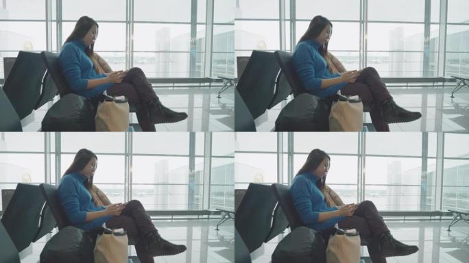 东南亚年轻女性旅行者，手持手机坐在慕尼黑国际机场的玻璃窗旁，滚动智能手机应用程序，检查航班