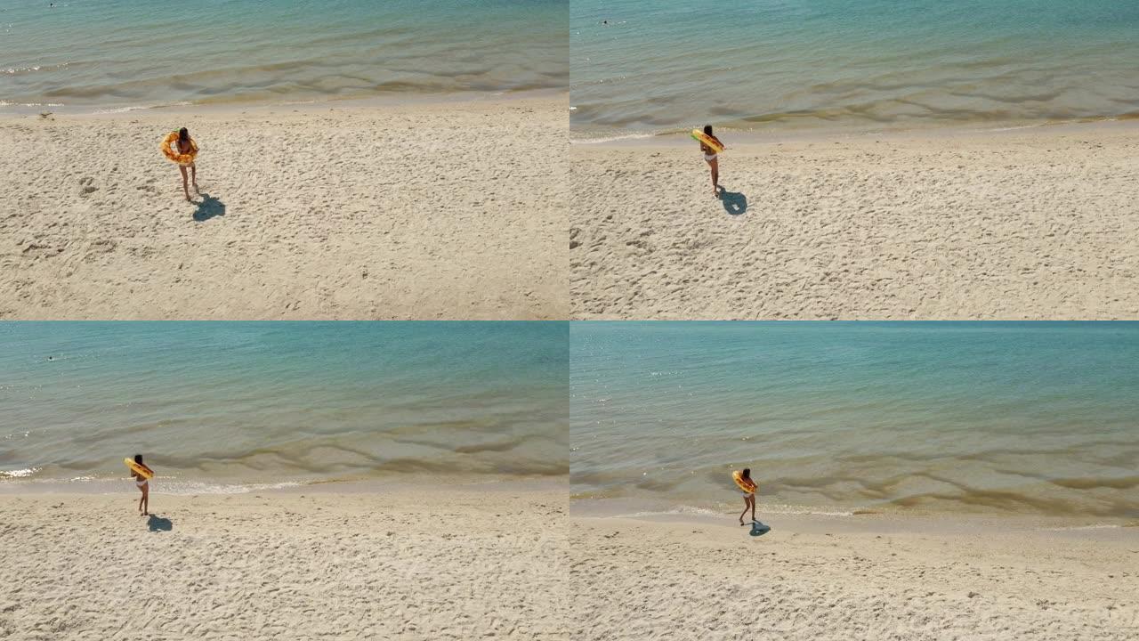 身材苗条的晒黑女人拿着巨大的漂浮菠萝戒指走在沙滩上的航拍画面。在热带海滩度假享受暑假的女孩。暑假和夏
