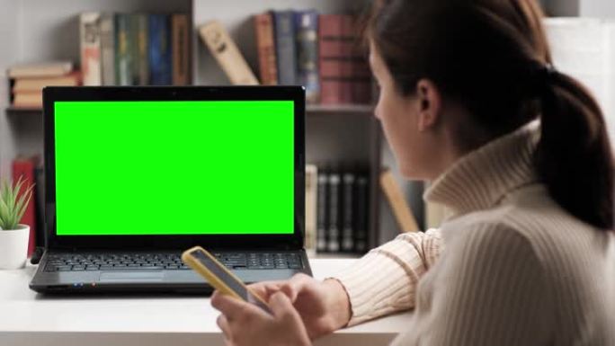 女人坐在桌子旁，看着绿屏笔记本电脑，她输入屏幕键盘并在internet上进行在线视频连接。色度键电脑
