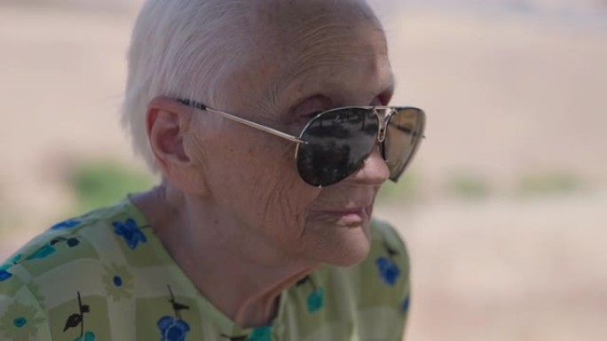 特写戴着墨镜的白发老妇人看着远处思考。阳光明媚的夏日，白种人体贴的退休人员在户外的爆头侧角视图。