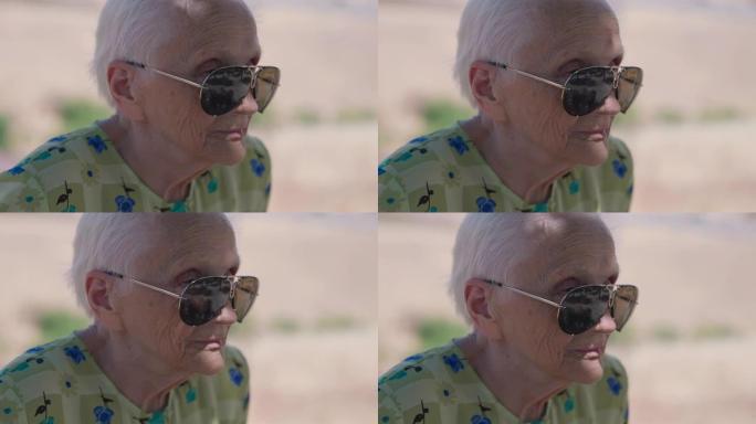 特写戴着墨镜的白发老妇人看着远处思考。阳光明媚的夏日，白种人体贴的退休人员在户外的爆头侧角视图。