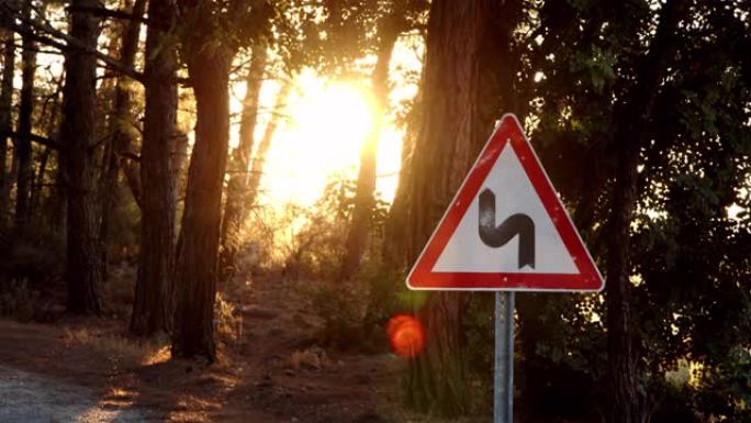 红色框架白色背景上蜿蜒的路标的近景。三角形标志，在日落或黎明时警告森林中的弯曲道路。