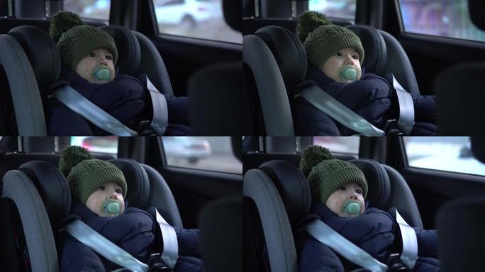 一个婴儿坐在汽车座椅上。儿童车内安全。冬天，一个男孩在窗户旁边的汽车上戴着扳机，戴着帽子。4k