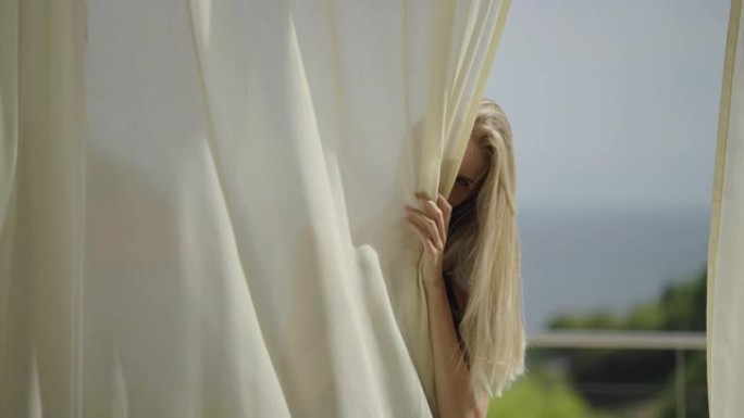 隐藏在透明窗帘后面的金色长发诱人的女人。与笼罩在神秘中的相机调情