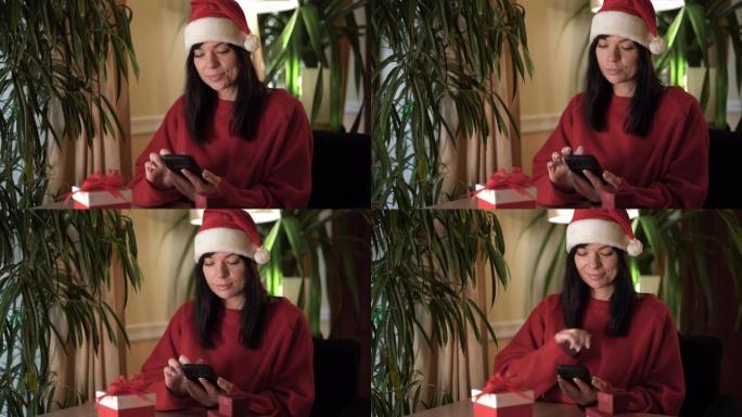 穿着圣诞老人服装的女人坐在家里的桌子旁，手里拿着智能手机，翻阅新闻。使用互联网与亲人交流的概念