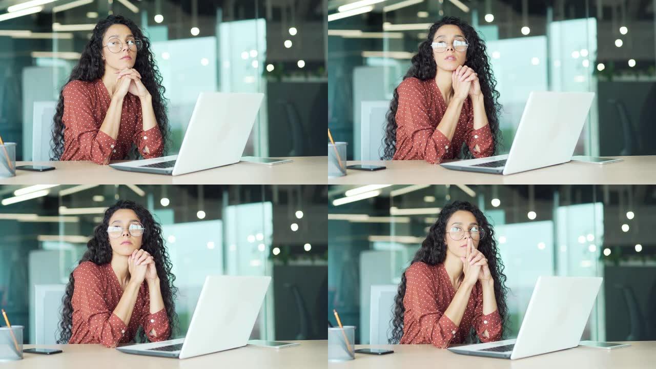 年轻严肃关心的西班牙裔女商人坐在办公桌前看着当代公司办公商务技术概念肖像中的笔记本电脑
