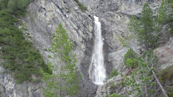 空中无人机的镜头缓慢地穿过树冠，飞向一个引人注目的瀑布，因为它在瑞士的一个陡峭的山崖边缘上瀑布。