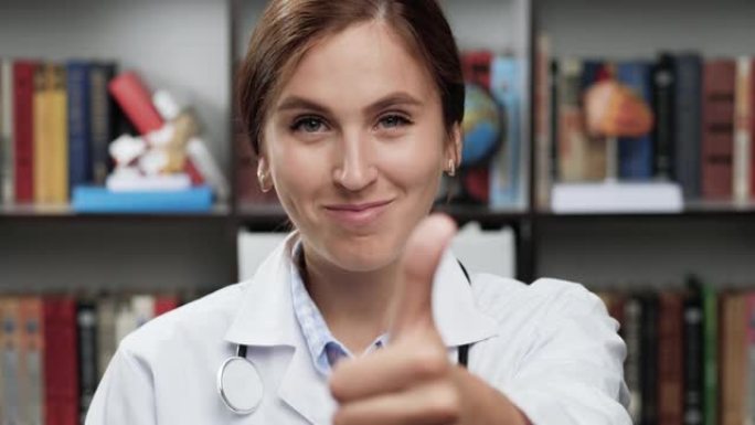 积极的医生竖起大拇指。穿着白大褂和听诊器的友好女医生看着相机，露出拇指，点头和微笑。爆头