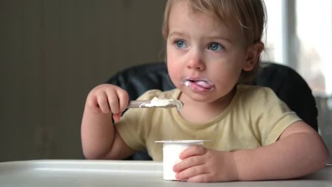 蹒跚学步的孩子坐在高脚椅上吃希腊酸奶。有趣可爱的宝宝在厨房吃饭。小女孩学习吃饭。孩子在家用硅胶勺子喂