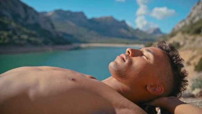 特写的年轻人躺在绿松石湖边的石头栅栏上晒日光浴