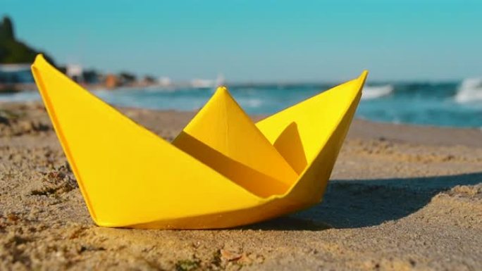 黄纸船在海边的沙滩上，在阳光明媚的日子里有白色泡沫的海浪背景下。旅游、航海、梦想、目标、决心、目的性