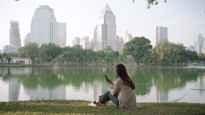 亚洲妇女很高兴通过早上在城市公园的电话在线聊天来放松身心，人们在城市自然环境中放松身心